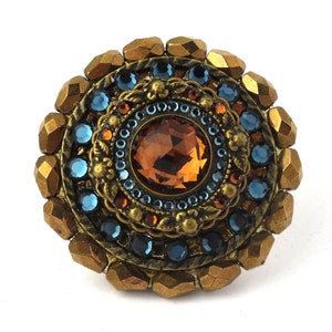 Peut inclure: Une bague ronde dorée avec une grande pierre facetée de couleur ambre entourée de petites pierres bleues. La bague est décorée de détails complexes et a un bord extérieur texturé.