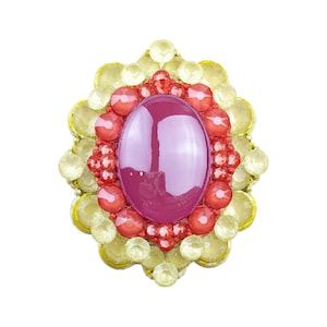 Puede incluir: Broche ovalado con una gran piedra central pulida de color magenta. El centro está rodeado por un anillo de cuentas rojas, que a su vez está rodeado por un borde de piedras en forma de pétalo de color amarillo pálido, todo sobre una base dorada.
