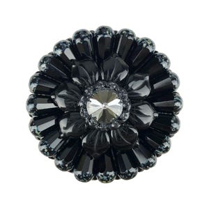 May include: A circular brooch featuring a central faceted clear crystal surrounded by multiple layers of black, petal-shaped beads. The outer beads have a speckled finish with small white and grey dots, creating a layered, flower-like appearance.