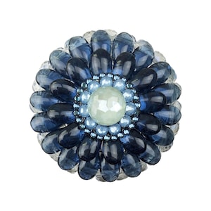 Op de afbeelding: Een ronde broche met een grote, gefacetteerde licht muntgroene middensteen, omringd door meerdere lagen donkerblauwe, doorschijnende, bloembladachtige glaskralen en een ring van kleinere, lichtblauwe iriserende kralen.