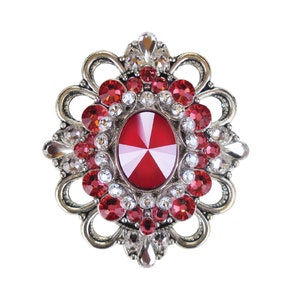 Könnte beinhalten: Eine verzierte Brosche mit einem ovalen, roten Mittelstein, umgeben von kleineren roten und klaren Kristallen. Die Brosche hat ein silberfarbenes Metall-Filigran-Design, das einen Vintage- oder Antik-Look erzeugt. Die Kristalle sind facettiert und reflektieren das Licht.
