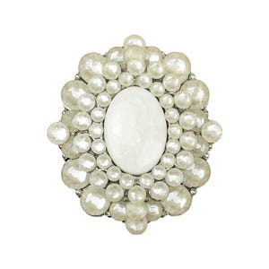 Puede incluir: Broche ovalada blanca con una piedra central y rodeada de perlas falsas de varios tamaños. El diseño es ornamentado y texturizado, con un engaste de metal plateado. Una pieza de joyería llamativa.
