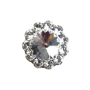 May include: A circular brooch featuring a large, faceted clear central stone surrounded by a halo of twelve smaller, round, brilliant-cut clear stones. The design is symmetrical, with the stones set in a metallic frame that reflects light.