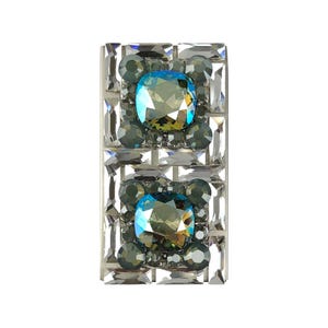 May include: Rectangular silver-toned accessory with a mosaic of clear and gray crystals. Two large, square-cut, iridescent blue-green stones are centered, surrounded by smaller gray crystals. The design is symmetrical and eye-catching.