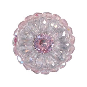 Pode incluir: Um broche circular com um cristal central redondo facetado em rosa, rodeado por um anel de contas de cristal transparentes e alongadas, com uma borda externa de contas de cristal menores, redondas e rosa claro sobre uma base prateada.