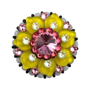 May include: A circular brooch featuring a large central pink crystal surrounded by a ring of smaller red crystals. The outer layer consists of yellow petals, each accented with a clear iridescent crystal, finished with a border of white beads.