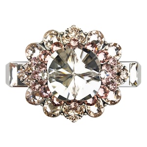 Puede incluir: Anillo plateado con un gran cristal transparente facetado en el centro, rodeado de cristales más pequeños, redondos, rosas y color champán. La banda del anillo tiene detalles rectangulares a cada lado del diseño floral.