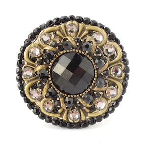 以下が含まれることがあります： 中央に黒いファセットカットの宝石がついた、丸い金色調のブローチ。ブローチは、黒と透明の小さなラインストーンで囲まれています。