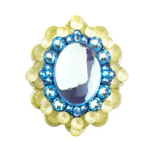 May include: An oval-shaped, ornate brooch with a large, iridescent center stone. The brooch is surrounded by a ring of blue gems, which are then encircled by a border of yellow-green gems. The design is symmetrical and eye-catching.