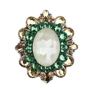 Puede incluir: Un broche ornamentado con una gran piedra central ovalada, facetada y blanquecina. El centro está rodeado por un anillo de pequeñas gemas verdes, y el borde exterior presenta gemas doradas y verdes alternadas, todo engastado en metal plateado.
