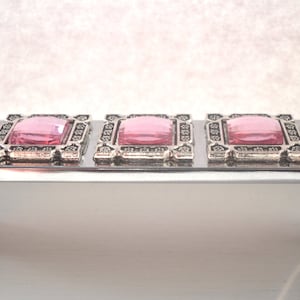 Könnte beinhalten: Ein silberner Schubladenzug mit fünf pinkfarbenen quadratischen Edelsteinen, die in verzierten silbernen Rahmen gefasst sind.