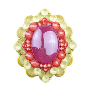 May include: An oval-shaped brooch with a large, polished, magenta-colored center stone. The center is surrounded by a ring of smaller, red-orange stones, and a border of yellow-toned, faceted gems. The brooch has a gold-colored metal setting.