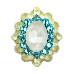 Puede incluir: Una joya ornamental con una piedra central ovalada, blanca y facetada. Está rodeada de piedras preciosas turquesas y amarillo pálido, dispuestas en un patrón floral o de estrella. El engaste parece plateado.