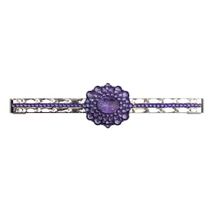Puede incluir: Horquilla plateada con una joya central ovalada de color púrpura rodeada de piedras púrpuras más pequeñas. La horquilla tiene un diseño lineal con cristales transparentes facetados y una fina línea púrpura.