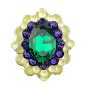 Puede incluir: Broche ovalado con una gran piedra central verde esmeralda facetada, rodeada de cristales más pequeños de color púrpura oscuro y amarillo pálido. El broche tiene un diseño floral, con los cristales amarillos formando el borde exterior.