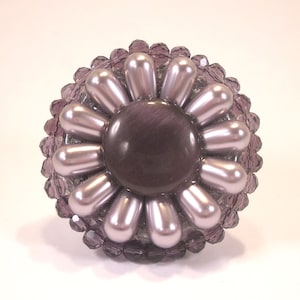 以下が含まれることがあります： 紫色の花の形をしたブローチ。大きな丸い紫色のセンターに、小さな真珠のような白いビーズのリングが囲まれています。ブローチの外縁は、小さな紫色のファセットカットのビーズでできています。