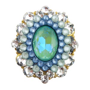 Puede incluir: Broche ovalada con una piedra central verde iridiscente, rodeada de cuentas azules y celeste. El borde exterior está adornado con cristales transparentes. El engaste tiene una base dorada. Accesorio decorativo.