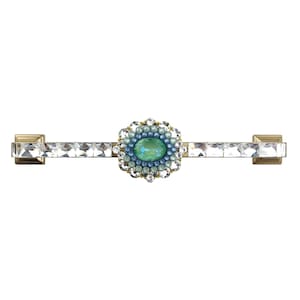 Könnte beinhalten: Ein dekorativer Schrankgriff mit rechteckiger Form. In der Mitte befindet sich ein ovaler, hellgrüner Edelstein, der von blauen und klaren Steinen umgeben ist. Der Griff ist mit klaren, rechteckigen Kristallen und goldfarbenem Metall akzentuiert.