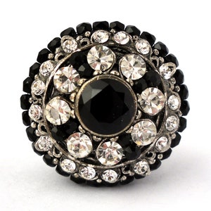 May include: A round, silver-toned ring with a central black gemstone surrounded by clear and black crystals. The ring has a detailed, ornate design, with a mix of large and small stones. The ring is set against a white background.