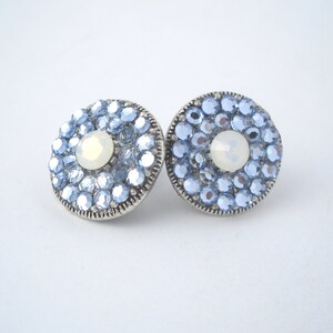 May include: A pair of silver stud earrings with a round design. Each earring features a cluster of light blue crystals surrounding a white opal-like stone in the center.