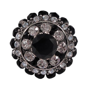 以下が含まれることがあります： 中央に大きな黒いラインストーン、周囲に小さなクリアなラインストーンが散りばめられた、黒と銀の丸いブローチです。