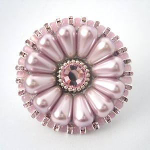 以下が含まれることがあります： 中央に大きなピンク色のクリスタル、周囲に小さなピンク色のパールと銀色のビーズがついた、ピンク色の花のブローチです。