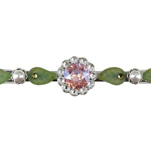Puede incluir: Una pinza para el pelo plateada con un centro de pedrería rosa rodeado de pedrería transparente. La pinza tiene dos acentos verdes en forma de lágrima a cada lado y dos acentos de perla blanca.