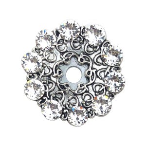 Puede incluir: Un botón redondo de metal plateado con un intrincado patrón de volutas en relieve y una abertura circular central. El borde exterior está adornado con diez cristales redondos, transparentes y facetados, dispuestos en círculo.