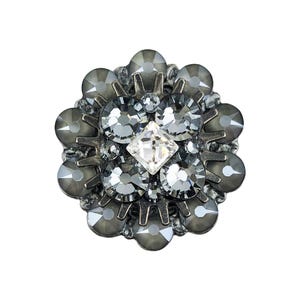 May include: A circular decorative button featuring a dark metallic base adorned with multiple faceted grey crystals arranged in a floral pattern, centered with a single clear diamond-shaped crystal, creating a textured and reflective surface.