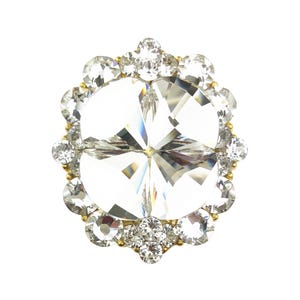 Peut inclure: Un grand bijou en cristal clair et facetté avec une monture en métal doré. Le bijou est entouré de cristaux plus petits et transparents, créant un design circulaire. Les cristaux scintillent sur fond blanc.