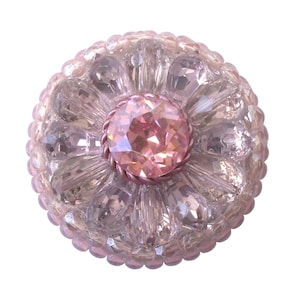 以下が含まれることがあります： 中央に大きなピンク色のラインストーンが付いたピンク色のガラスボタン。ボタンは小さなラインストーンで囲まれており、隆起したテクスチャのあるデザインになっています。
