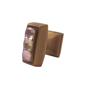 Puede incluir: Un pomo de armario de latón con una base rectangular y una parte superior cuadrada. El pomo tiene tres cristales de vidrio rosa y transparente incrustados en la parte delantera.