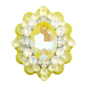 Könnte beinhalten: Ein dekoratives Strass-Ornament in Gelb und Weiß. In der Mitte befindet sich ein großer, oval geformter gelber Kristall, umgeben von kleineren weißen und gelben Steinen. Der äußere Rand hat ein gewelltes Design.