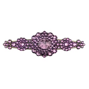 Puede incluir: Horquilla decorativa con una gema central ovalada de color púrpura, rodeada de piedras más pequeñas a juego. La horquilla tiene un diseño floral con racimos adicionales de piedras púrpuras a cada lado, sobre una base de metal dorado.