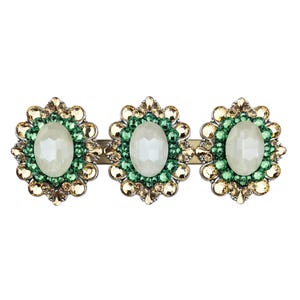 Op de afbeelding: Decoratieve haarclip met drie bloemvormige versieringen. Elke bloem heeft een centrale ovale, gebroken witte edelsteen, omringd door groene en goudkleurige kristallen. De clip is goudkleurig.