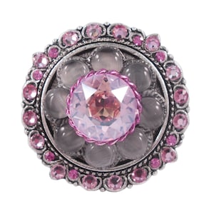 以下が含まれることがあります： 中央に大きなピンクのラインストーン、周囲に小さなラインストーンと銀の花柄が施された、銀とピンクのジュエリーブローチです。