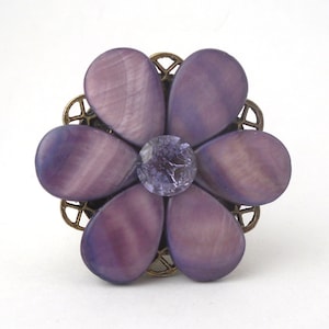 Puede incluir: Un anillo de flor púrpura con un centro de cristal transparente. La flor está hecha de concha iridiscente y está rodeada por un diseño de filigrana de bronce.