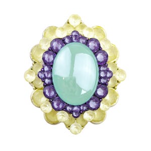 May include: A decorative brooch featuring a large, oval, pale green gemstone at the center, surrounded by a ring of purple gems and a border of yellow gems. The brooch has a silver-colored metal setting.