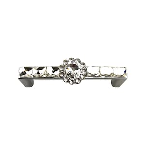 May include: A silver-toned metal cabinet pull featuring a horizontal bar adorned with a row of square clear crystals and a central circular floral-shaped cluster of smaller clear crystals. The hardware has two mounting points on the ends.