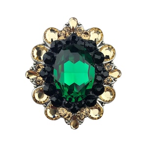 Puede incluir: Un broche ornamentado con una gran gema ovalada verde esmeralda en el centro, rodeada de pequeños cristales negros y amarillo dorado. El broche tiene un engaste de metal plateado y un diseño detallado, similar a una flor, creando una apariencia lujosa.