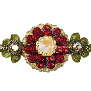 Könnte beinhalten: Eine goldfarbene Brosche mit einem roten und goldenen Blumendesign. Die Blume ist von grünen Blättern umgeben und hat einen klaren Kristall in der Mitte.