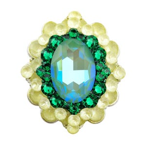 May include: An oval-shaped, jewel-toned brooch with a central, iridescent, teal-colored stone. The stone is surrounded by a ring of smaller, emerald-green gems, and a border of pale yellow, rounded stones. The brooch has a metallic base.