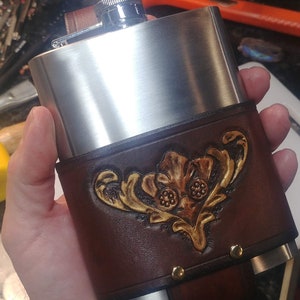 May include: A stainless steel flask with a brown leather carrying case. The case has a gold embossed design of a stylized flower.