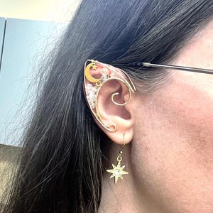 May include: Gold ear cuff with a crescent moon and star design, accented with clear crystals. A matching gold star dangle earring is also visible.