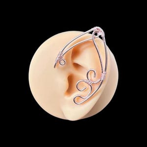 May include: A rose gold ear cuff with a delicate, intricate design. The cuff is made of wire and has a curved shape that wraps around the ear.