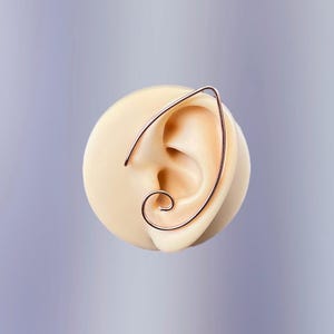 May include: A rose gold ear cuff is displayed on a light-colored ear model. The ear cuff wraps around the ear, with a curved design that includes a spiral at the bottom. The background is a soft, gradient purple.