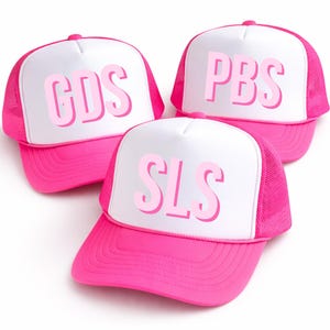 May include: Three pink trucker hats with white front panels. Each hat features a different set of pink capital letters: "CDS", "PBS", and "SLS". The hats have a mesh back and a curved brim.