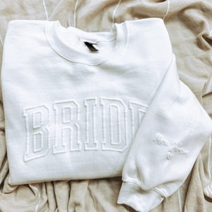 Peut inclure: Sweat-shirt blanc avec le mot "BRIDE" brodé en gros caractères gras. Le sweat-shirt a également la phrase "Mrs. to be" brodée sur la manche.