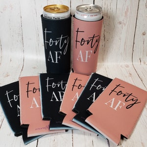 Puede incluir: Un conjunto de ocho enfriadores de latas negros y rosados con el texto "Forty AF" impreso en ellos. Los enfriadores est&aacute;n dispuestos en semic&iacute;rculo sobre una superficie de madera.