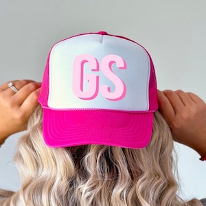 May include: A hot pink trucker hat with a white front panel featuring the letters "GS" in a layered pink font. The hat has a mesh back and a curved brim. The hat is being worn.